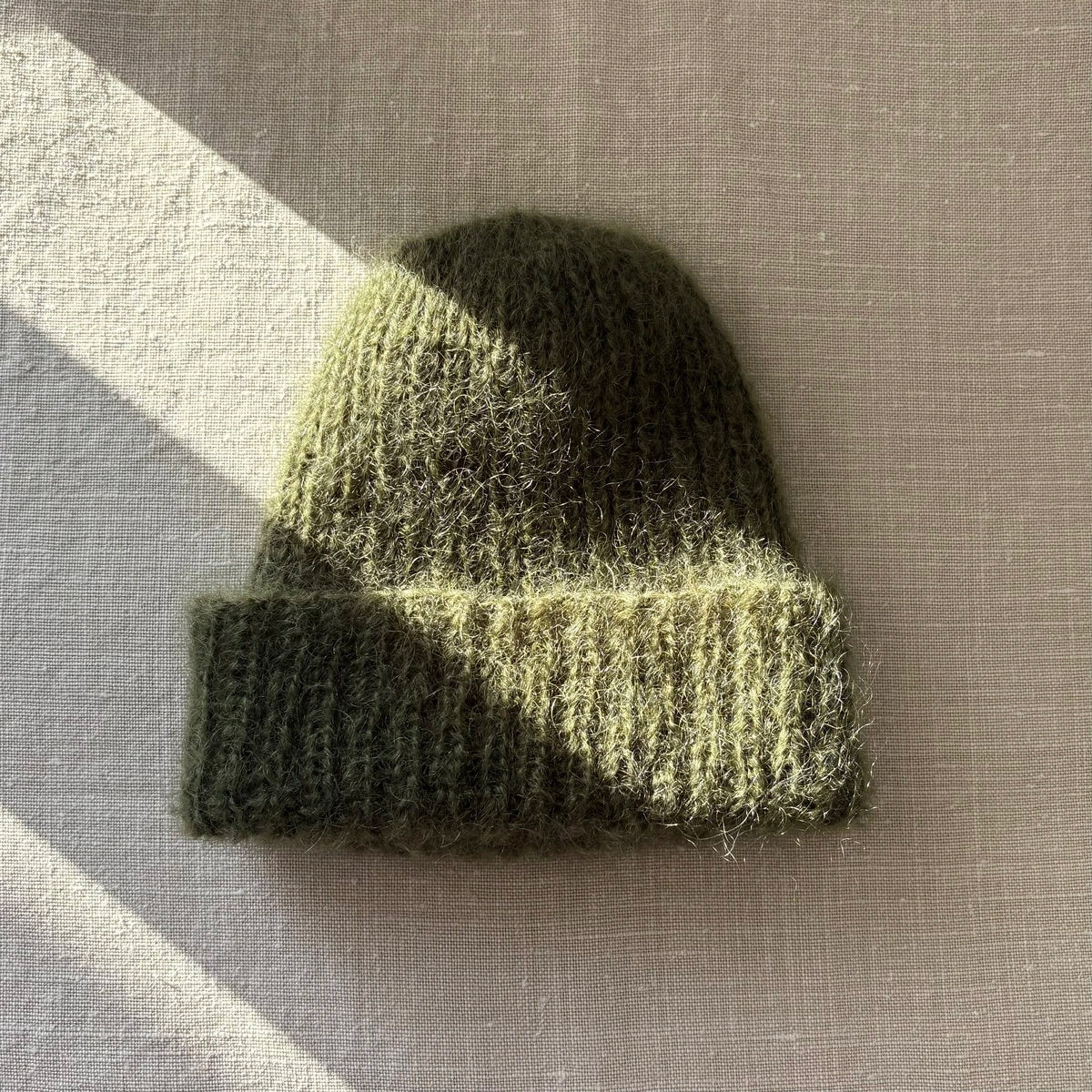 bonnet en mohair vert kaki posé sur du lin avec un rayon de soleil