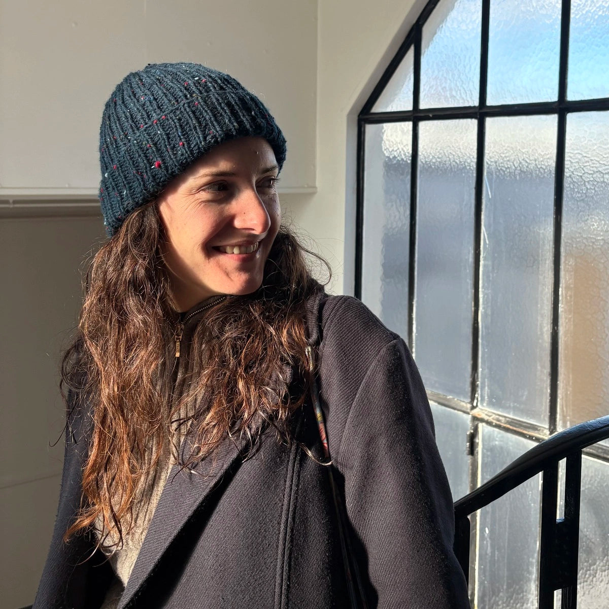 femme aux cheveux longs dans un hall d'escalier souriant et portant un bonnet en tweed bleu marine