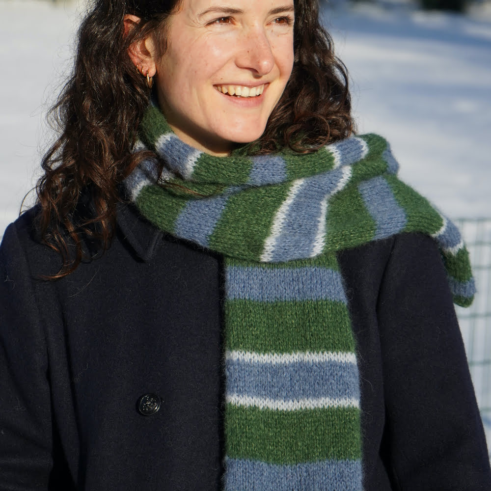 femme brune souriant au soleil avec un caban et une écharpe rayée bleu et verte tricotée