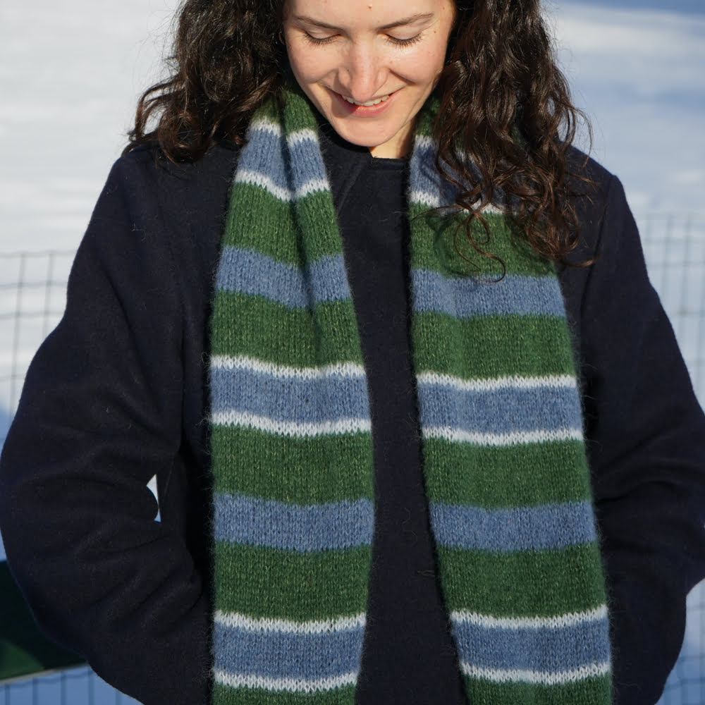 femme brune baissant la tête et souriant, regardant une écharpe rayée bleu et verte tricotée